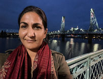 Nahal is standing on a bridge with a big city scape behind her. She has brown hair pulled back and is wearing a red scarf
